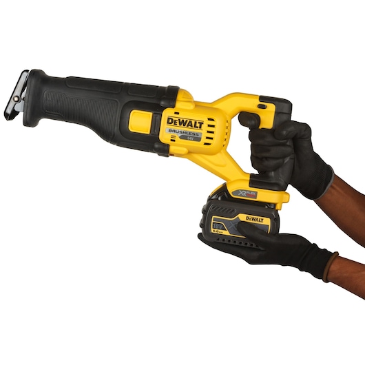 A person wearing black gloves is holding a yellow and black DEWALT cordless reciprocating saw. The saw features a brushless motor and an 18V XR FLEXVOLT battery. The product is shown against a white background.