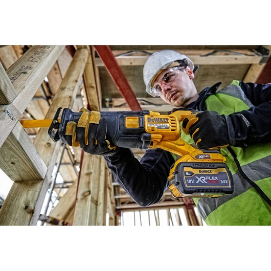 A person wearing safety gear uses a DEWALT cordless reciprocating saw, model DCS389N, to cut through wooden beams at a construction site. The tool is powered by an 18V/54V XR FLEXVOLT lithium-ion battery.