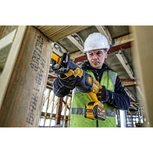 A person wearing safety gear operates a DEWALT DCS389N cordless reciprocating saw on a construction site. The tool is yellow and black, and the person is cutting through a wooden beam.