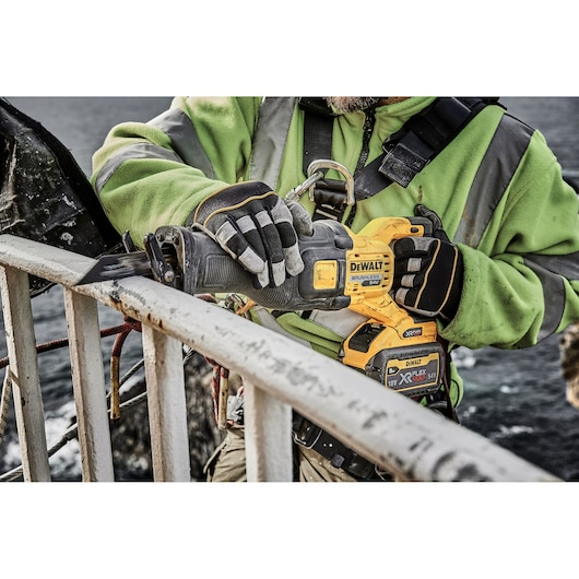 A person wearing a green safety jacket and gloves is using a DEWALT DCS389X2 cordless reciprocating saw to cut through a metal railing outdoors.