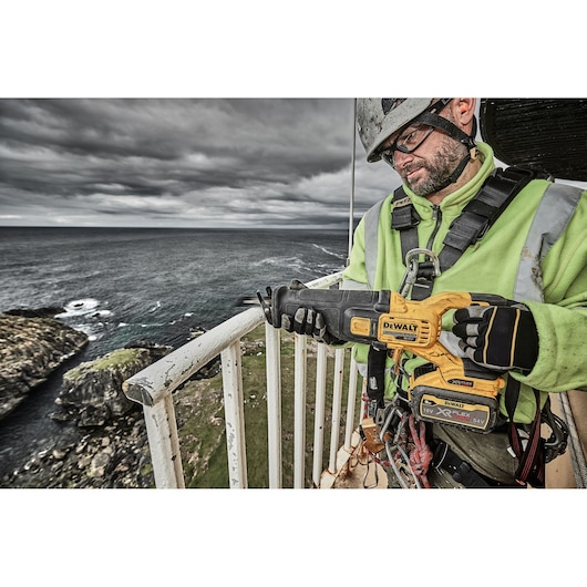 A worker in safety gear is holding a DEWALT cordless reciprocating saw (model DCS389X2) on an outdoor balcony overlooking the sea and rocky coastline. The worker's face is obscured.