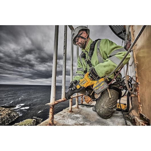 A worker wearing a safety helmet and high-visibility jacket uses a DEWALT DCS389X2 cordless reciprocating saw to cut through a rusty metal railing outdoors near the coast, with dark clouds overhead.