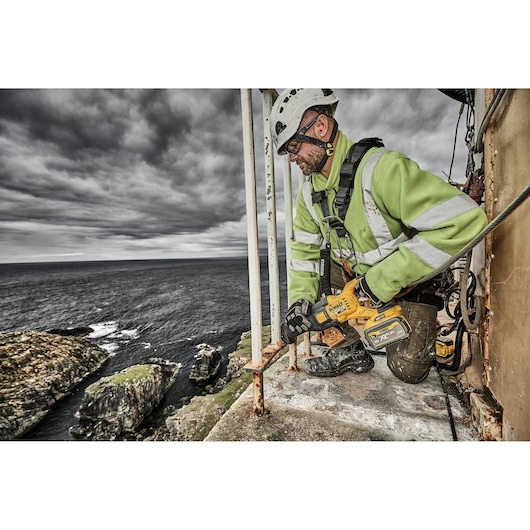 A person wearing safety gear and a green reflective jacket is using a DEWALT cordless power tool to cut metal rods on an outdoor platform near the edge of a cliff, with an ocean and rocky coastline in the background.