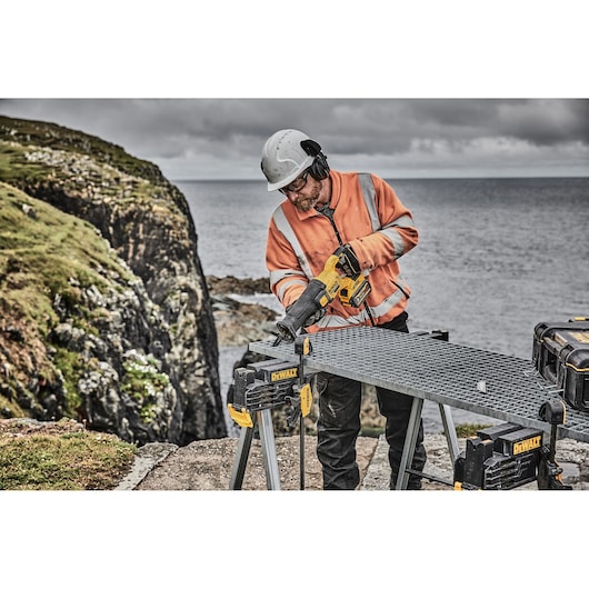 A worker wearing a helmet and an orange safety jacket is using a DEWALT cordless reciprocating saw on a metal workbench outdoors, near a rocky coastline.