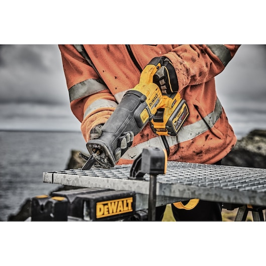 A person wearing an orange work jacket is using a DEWALT cordless reciprocating saw to cut through a metal grate outdoors. The DEWALT logo is visible on the saw and workbench.