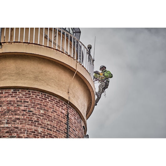 A worker equipped with safety gear is using a DEWALT power tool while suspended by ropes on the side of a brick and concrete structure, possibly performing maintenance or construction.