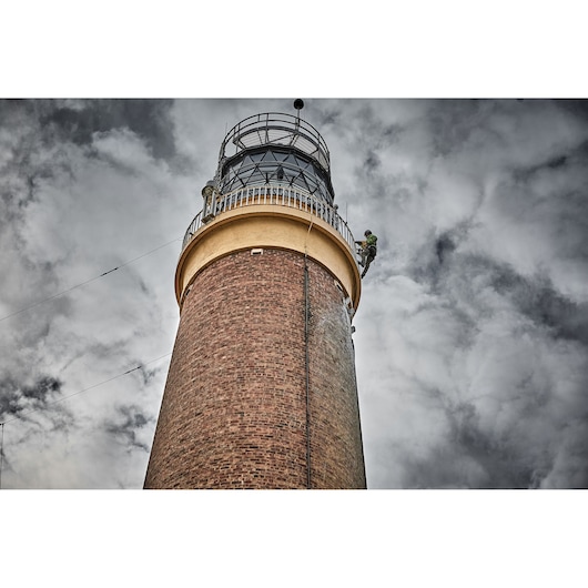 A tall brick lighthouse viewed from below against a cloudy sky, with a worker wearing safety gear ascending near the top of the structure.
