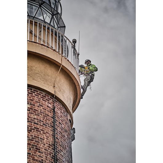 A person wearing safety gear is suspended on ropes, working on the exterior of a brick lighthouse with a railing near the top. The sky is cloudy.