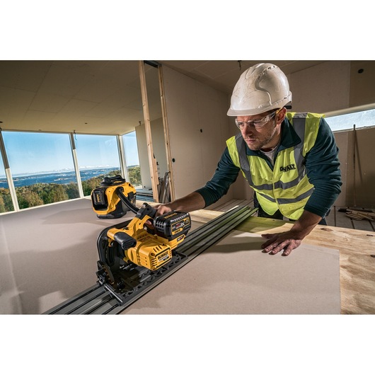 Worker uses a DEWALT DCS520NT saw to cut sheet material indoors, wearing safety gear and high-visibility vest.