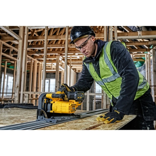 A worker using a DEWALT DCS520T2-XE cordless track saw in a construction site with wooden framing in the background. The worker is wearing a safety vest, gloves, and a hard hat.