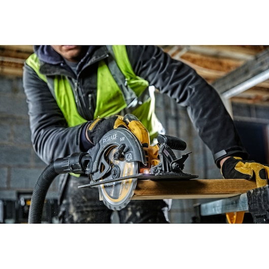 A person wearing a yellow safety vest and gloves is using a DEWALT DCS575NT 54V cordless circular saw to cut a piece of wood in a construction site.