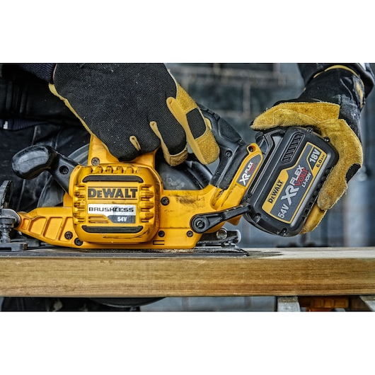 A close-up view of a DEWALT DCS575NT brushless 54V circular saw being held by a person wearing black and yellow protective gloves, shown working on a piece of wood.