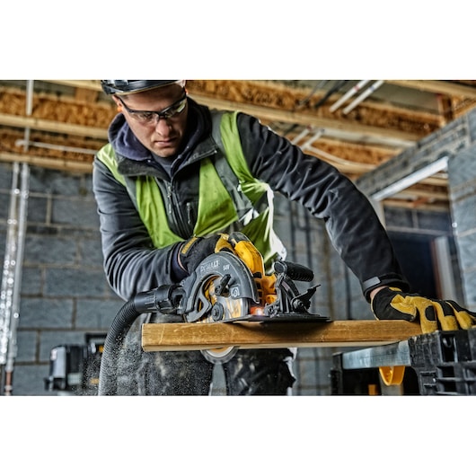 Person uses a DEWALT circular saw to cut wood on a construction site, wearing safety gear and gloves.