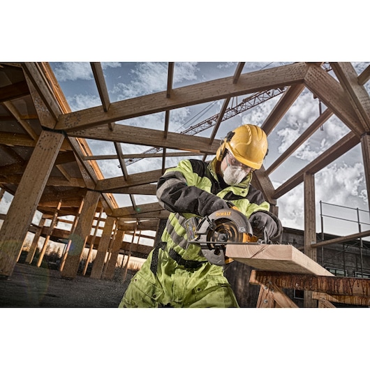 A construction worker wearing safety gear uses a DEWALT circular saw to cut wood at a building site. The worker is outdoors, and wooden framing structures are visible in the background.