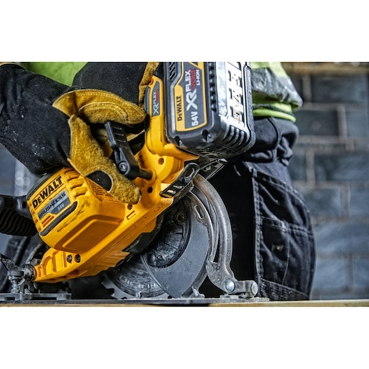 Close-up of a person wearing gloves operating a DEWALT cordless circular saw with a 54V XR FLEXVOLT battery, cutting through wood in a workshop setting.