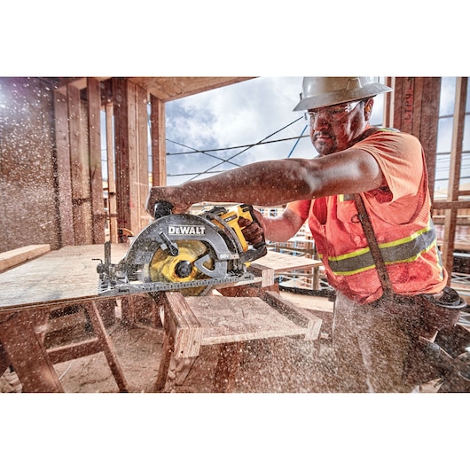 A person wearing a safety helmet and orange reflective vest is using a DEWALT DCS577B cordless circular saw to cut wood at a construction site, with sawdust flying in the air.