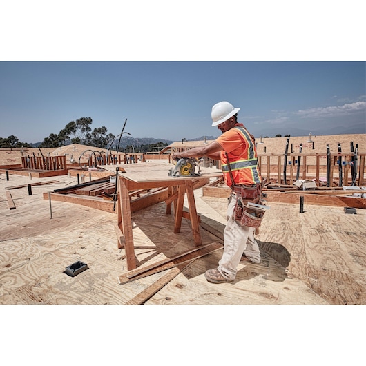 A construction worker wearing a hard hat and safety vest is using a DEWALT circular saw to cut plywood on a building site. The worker stands next to a sawhorse with tools attached to their belt and the site shows ongoing construction with wood and pipes in the background.