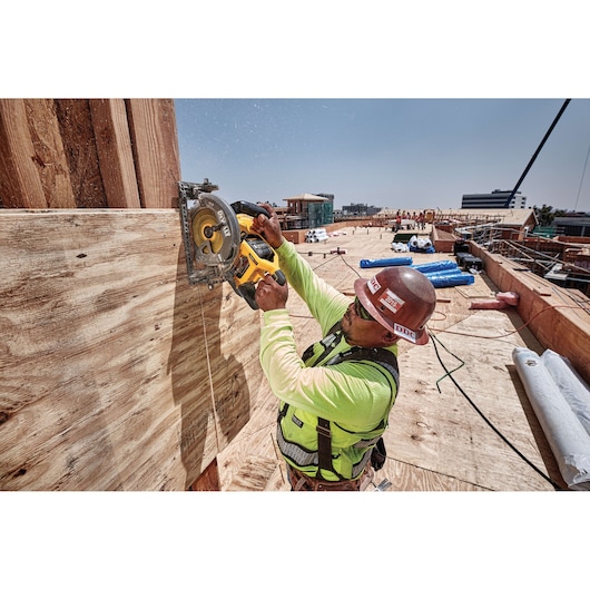 A construction worker wearing a safety helmet and vest uses a DEWALT circular saw to cut plywood on a jobsite. The worker is positioned outdoors, surrounded by building materials and other constructions under a clear sky.