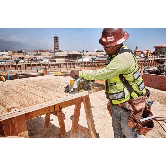 A construction worker wearing a safety vest and hard hat operates a DEWALT DCS577B circular saw to cut plywood on a jobsite, with buildings and scaffolding visible in the background.