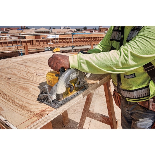 A person wearing a safety vest and long-sleeved shirt is using a DEWALT DCS577B cordless circular saw to cut a sheet of plywood at a construction site.
