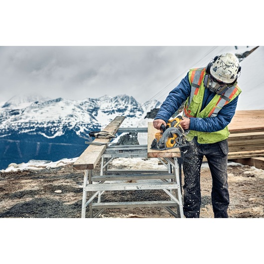 A construction worker wearing a helmet and safety vest uses a DEWALT circular saw to cut wood outdoors at a snowy mountain site.