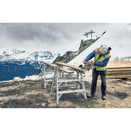 A construction worker wearing safety gear uses a DEWALT DCS577B circular saw to cut a wooden plank on a jobsite set in a snowy mountain environment. The plank is supported by two metal sawhorses, and there are snow-covered peaks and ski lift structures in the background.