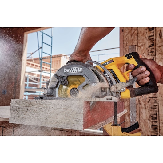 A close-up of a person using a DEWALT DCS577B cordless circular saw to cut through a piece of wood at a construction site. Sawdust is flying as the saw operates.