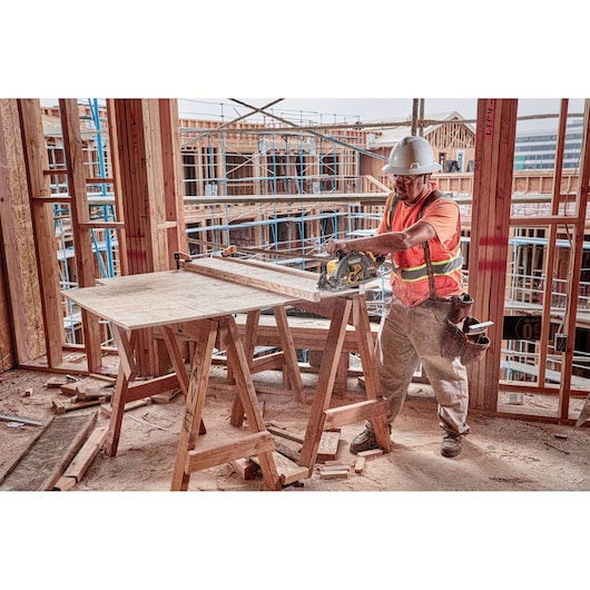 A construction worker wearing a hard hat and safety vest uses a DEWALT circular saw to cut wood on a work table at a building site with partially constructed wooden framework in the background.