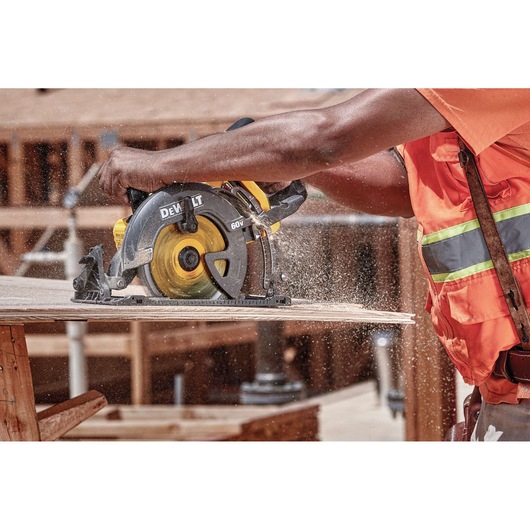 A construction worker wearing an orange safety vest uses a DEWALT DCS577B cordless circular saw to cut a piece of wood, with sawdust flying in a workshop or construction site.