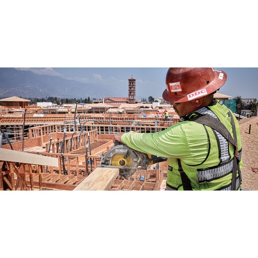 A construction worker wearing a safety vest and helmet is using a DEWALT DCS577B circular saw to cut a wooden beam at a building site, with unfinished wooden structures and a tower in the background.
