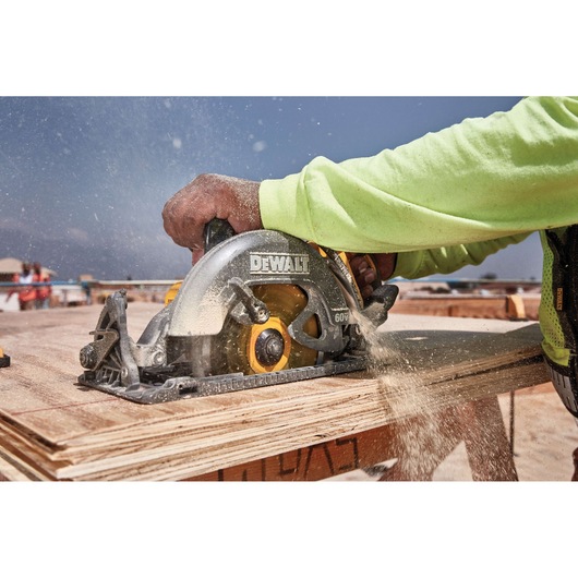 A person in a neon green long-sleeve shirt uses a DEWALT cordless circular saw to cut through wooden boards at an outdoor construction site, with sawdust spraying from the blade.