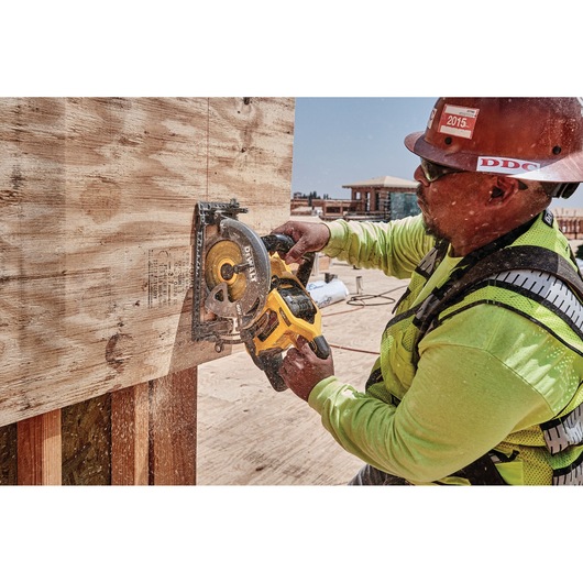 A worker using a DEWALT DCS577B cordless circular saw to cut plywood on a construction site. The worker is wearing safety gear, including a hard hat, a high-visibility vest, and gloves.