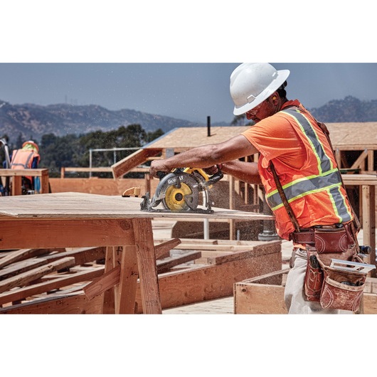 A construction worker wearing a safety vest and helmet is using a DEWALT circular saw (model DCS577B) to cut wood at an outdoor building site. The worker’s face is blurred for privacy and sawdust is flying as the tool operates.