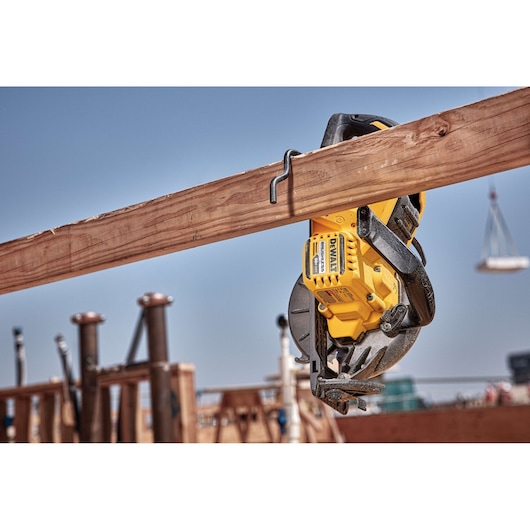 A DEWALT DCS577B cordless circular saw hanging on a wooden beam at a construction site, with clear sky in the background.
