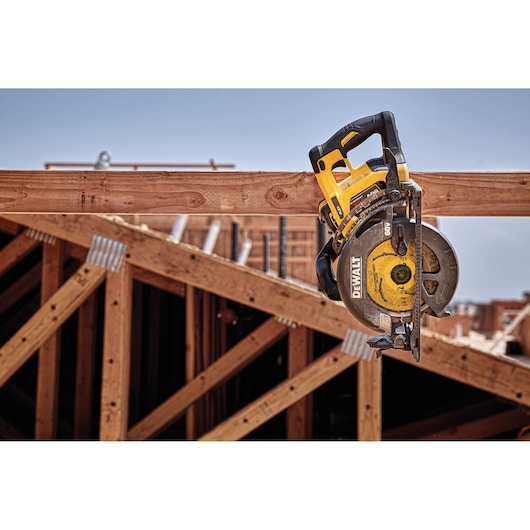 A DEWALT cordless circular saw is hanging from a wooden beam at a construction site, with wooden framing and structures visible in the background.