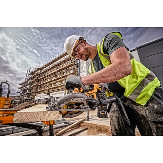 A construction worker wearing a safety vest, gloves, and a helmet is using a DEWALT circular saw (model DCS577N) to cut a wooden plank outdoors at a building site with scaffolding in the background.