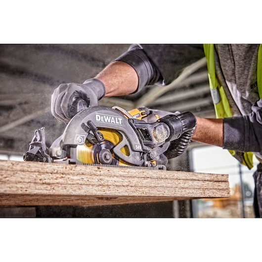 A close-up of a person using a DEWALT circular saw to cut a piece of wood in a construction setting. The person is wearing gloves and a safety vest.