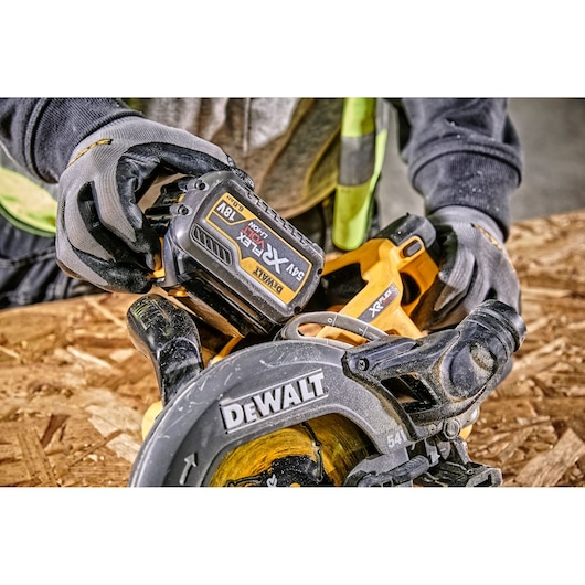 Close-up of a person wearing gloves and a safety vest using a DEWALT cordless circular saw on a wooden surface. The saw features a visible 54V/18V XR FLEXVOLT battery.