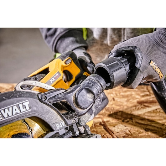 Close-up view of a person wearing gloves connecting a hose to a DEWALT cordless circular saw. The saw is being used on a piece of plywood.
