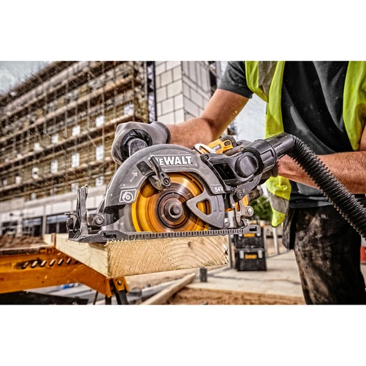 A DEWALT circular saw (model DCS577N) being used by a person wearing a safety vest and gloves to cut a piece of wood at a construction site outdoors.