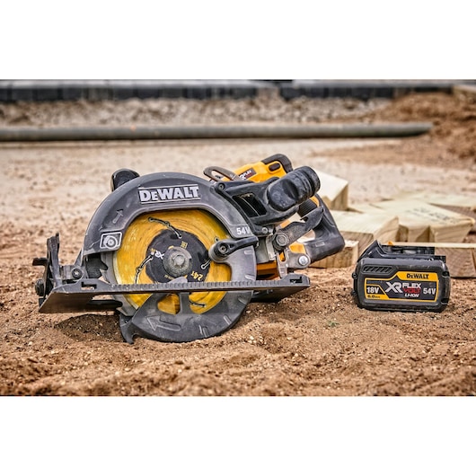 DEWALT cordless circular saw with a yellow blade and XR FLEXVOLT 18V/54V lithium-ion battery placed on sandy ground at a construction site, with wooden planks in the background.