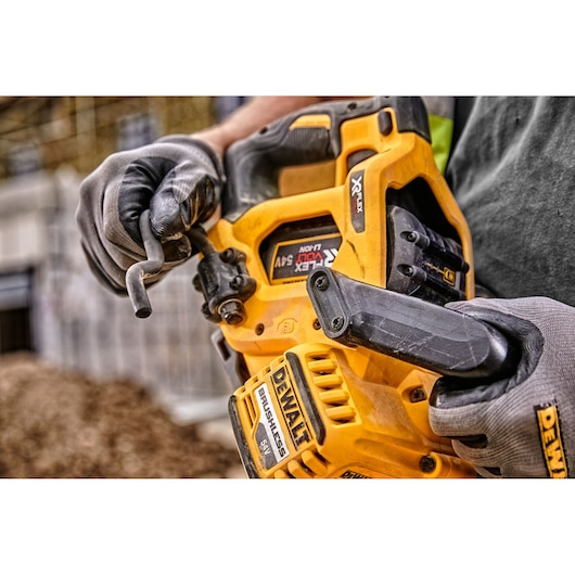 Close-up of a person wearing gloves holding a DEWALT cordless power tool, specifically a yellow and black brushless saw, with visible battery and adjustment wrench. The background shows an outdoor construction site.