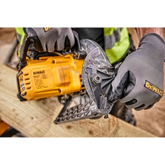 Close-up of gloved hands adjusting the angle on a yellow DEWALT DCS577N circular saw, positioned on a wooden plank at a construction site.