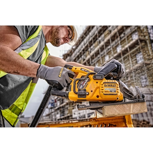 A person wearing protective gloves and a high-visibility vest is using a DEWALT DCS577N cordless circular saw to cut a piece of wood at a construction site. Sawdust is visible flying from the cut.