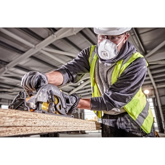 A worker wearing gloves and a safety vest uses a DEWALT DCS577N circular saw to cut wooden boards inside a construction site.
