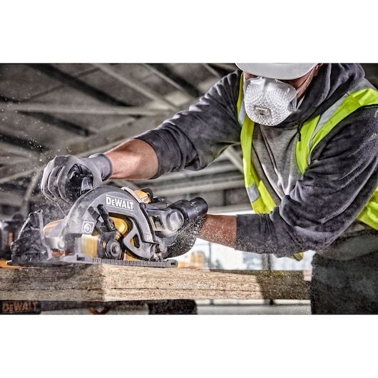 A person wearing safety gear, including a mask, gloves, and a high-visibility vest, is using a DEWALT circular saw to cut through a wooden plank, creating a cloud of sawdust in a construction setting.