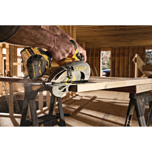 A person using a DEWALT DCS578X1 cordless circular saw to cut a wooden plank in an unfinished construction site.