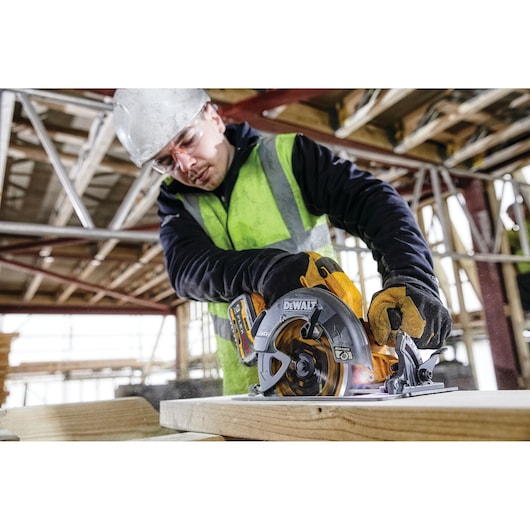 A person wearing a safety vest and gloves is using a DEWALT DCS578X2 circular saw to cut a piece of wood at a construction site. The face of the person is blurred for privacy.