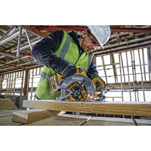 A person wearing a safety vest and helmet is using a DEWALT cordless circular saw (model DCS578X2) to cut wood on a construction site.