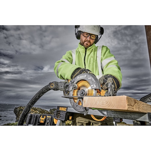 A person wearing safety gear operates a DEWALT DCS578X2 circular saw, cutting a piece of wood outdoors. The saw is connected to a vacuum hose, and the worker is dressed in a high-visibility jacket.
